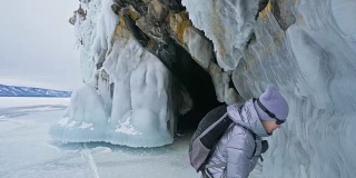 女人在贝加尔湖冰面上的旅行。冬岛之旅。女孩在冰岩石下行走。游客看着美丽的冰洞。极限跋涉和步行。背包客在大自然中休憩。
