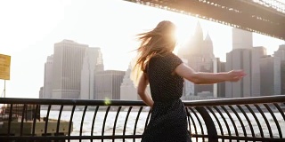 美丽兴奋的旅行者女孩在布鲁克林大桥的河堤上旋转，手臂在空中举起