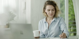 年轻女子用笔记本电脑工作，在办公室喝咖啡