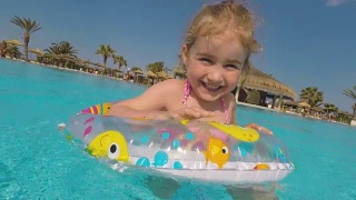 在炎热的夏日里，快乐的小女孩在室外游泳池里玩五彩缤纷的充气圈。孩子们在热带度假胜地玩耍。孩子们学游泳。家庭海滩度假。视频素材模板下载