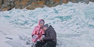 年轻的夫妇有乐趣在冬季散步的背景冰冻结的湖。情侣们坐在冰上，亲吻拥抱。快乐的年轻人在冰雪覆盖的冰面上漫步。蜜月。爱情故事。