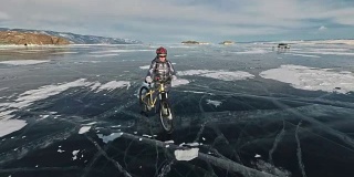在冰上，一个女人在自行车旁边行走。这个女孩穿着银色的羽绒服，背着背包，戴着头盔。结冰的贝加尔湖的冰。自行车的轮胎上覆盖着特殊的尖钉。