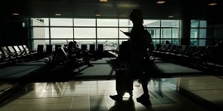 一位女士提着带轮子的包，手里拿着手机正在机场候机楼里走着。大窗户映衬下的剪影
