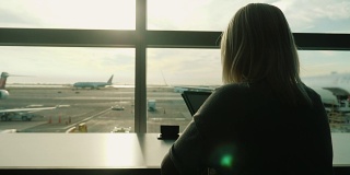 一位女士坐在机场候机楼窗口的桌边，享受着平板电脑。永远在线，旅行舒适
