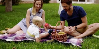 在一个阳光明媚的日子里，快乐的一家人坐在草地上野餐。他们的儿子就坐在附近。享受野餐。在户外