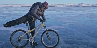 男人和他的自行车在冰上。骑自行车的人穿着灰色羽绒服，背着背包，戴着头盔。结冰的贝加尔湖的冰。自行车的轮胎上覆盖着特殊的尖钉。