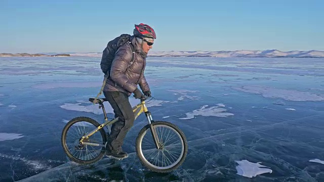 男人在冰上骑自行车。骑自行车的人穿着灰色羽绒服，背着背包，戴着头盔。结冰的贝加尔湖的冰。自行车的轮胎上覆盖着特殊的尖钉。