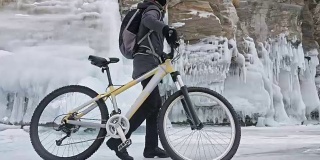 在冰洞附近，一个人在自行车旁边行走。有冰洞和冰柱的岩石非常漂亮。骑自行车的人穿着灰色羽绒服，背着背包，戴着头盔。旅行者正在骑自行车。