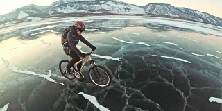 男人在冰上骑自行车。骑自行车的人穿着灰色羽绒服，背着背包，戴着头盔。结冰的贝加尔湖的冰。自行车的轮胎上覆盖着特殊的尖钉。
