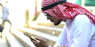 紧张的胡子阿拉伯商人坐在人行道上玩手机。