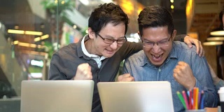 4K视频的亚洲商人在休闲西装工作笔记本电脑在现代的工作场所或咖啡店与同事，而他的成功来了，并祝贺一起，商业和自由职业者的概念