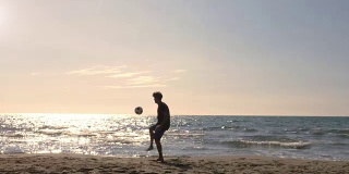 年轻男子在游泳裤做特技运球弹跳足球在海边在日落的海滩剪影