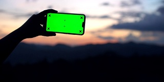 男人拿着智能手机的绿色屏幕与自然戏剧性的天空为背景，色度键，景观模式