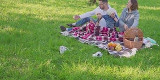 兴高采烈的一家人坐在公园的草地上野餐，都在吃早饭。有一个盛饭的篮子。新鲜的水果