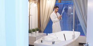 年轻有魅力的男人在一个豪华水疗中心休息白色长袍。一名男子站在全景窗口边讲手机