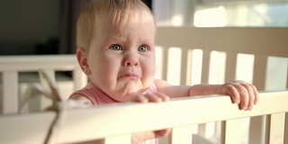 可怜的婴儿在家里的婴儿床上哭。不开心的小孩站在婴儿床里