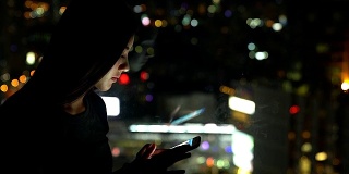 年轻的亚洲女性在夜晚使用手机，以城市的灯光为背景。