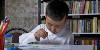 年轻专注的亚洲孩子在纸上画画，坐在桌子后面，男孩做作业，书架的背景