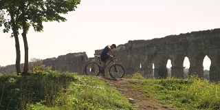 骑自行车的人在雄伟的古罗马渡槽前的土路上骑着自行车上山。年轻有吸引力的运动员与蓝色t恤和短裤运动装在parco degli acququedotti在罗马