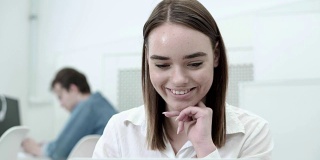 美丽的女学生在笔记本电脑屏幕上微笑阅读，她的同学在后台使用手机