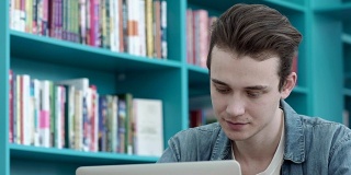 在大学图书馆使用笔记本电脑的学生。年轻男子在书架间做研究的特写