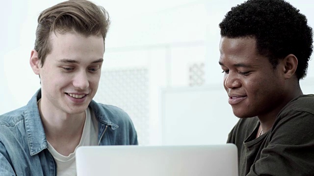 两个不同的男同学一起在笔记本电脑上做项目。年轻人看着屏幕，愉快地交谈着