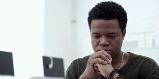 一个深思熟虑的非裔美国学生双手托着下巴在笔记本电脑屏幕上阅读。严肃的年轻人专注于工作