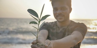 一个微笑的年轻人，双手捧着一株绿色的小植物或样本，对着冉冉升起的金色太阳