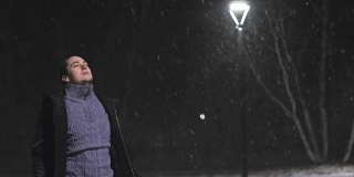 路灯下的那个人。在灯光的映衬下，正在下雪。慢动作，每秒180帧。雪花很漂亮。那个男人穿着一件凉爽的黑外套。