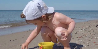 金发小男孩在海边玩耍。孩子在海边的沙滩上走着，玩着沙子和玩具。
