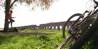 骑自行车的人站在一棵树下的岩石上，在古罗马的渡槽前看地图。年轻有吸引力的运动员与橙色运动装和背包parco degli acququedotti在罗马