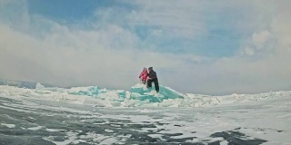 年轻的夫妇有乐趣在冬季散步的背景冰冻结的湖。情侣们坐在巨大的蓝色浮冰上，亲吻拥抱，从热水瓶里喝着茶。爱情故事。