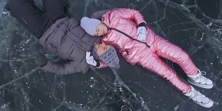 年轻的夫妇有乐趣在冬季散步的背景冰冻结的湖。情侣们躺在有裂缝的透明冰面上，嬉戏、亲吻、拥抱。从以上观点。蜜月。爱情故事。