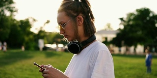 一个穿着白色T恤的年轻女孩在她的手机上打字的侧视图。她脖子上戴着黑色耳机。花时间在绿色公园。阳光照在背景上
