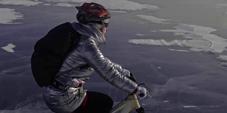 一个女人在冰上骑自行车。这个女孩穿着银色的羽绒服，背着自行车背包，戴着头盔。用四旋翼无人机拍摄。自行车的轮胎上布满了钉子。