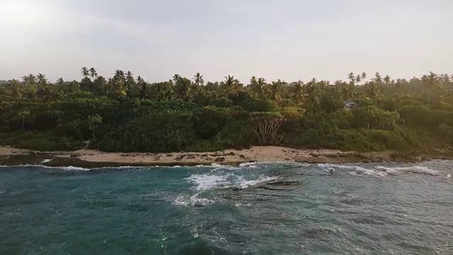 航拍镜头跟随汹涌的海浪涌向海岸，展现了一个充满热带树木、阳光明媚的异国小岛