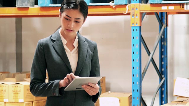年轻的女工在工厂或仓库的货架之间行走和检查库存。女人在仓库里检查用品并使用平板电脑。