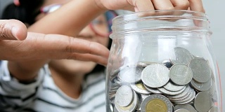 亚洲小女孩将硬币放入一个浅景深的玻璃罐中，选择焦点放在罐中