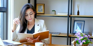 4K视频，美丽的亚洲商务女性坐在电脑笔记本前，阅读和翻动笔记本页与思考的脸在工作空间早上，商务人士的生活方式。
