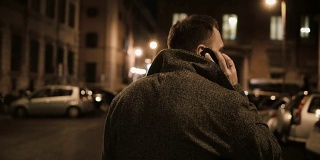 年轻英俊的男子走在晚上的街道上，使用智能手机，与朋友聊天的背影