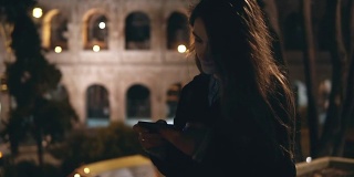 缓慢的运动。年轻漂亮的女人在意大利罗马的罗马斗牛场附近散步，晚上还在用智能手机