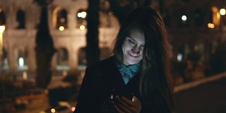 缓慢的运动。一名年轻的黑发女子晚上在意大利罗马斗牛场(Colosseum)附近散步，她正在使用智能手机