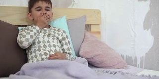生病的小男孩坐在床上，用毯子盖住自己，咳嗽