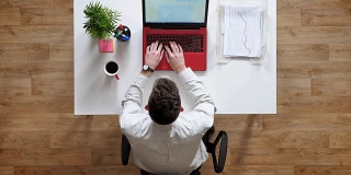 一个年轻人在笔记本电脑上打字，看着屏幕，坐在桌子后面拿着文件和咖啡