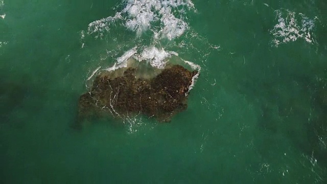 无人机锁定拍摄到透明的绿色海浪冲刷着海岸附近美丽海洋中的大石头，溅起泡沫