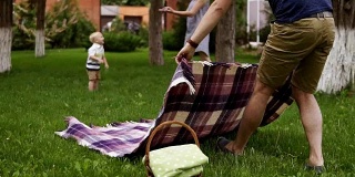 父亲穿着棕色短裤和蓝色衬衫正在铺开格子布准备野餐。在模糊的背景上，快乐的妈妈正在和她的小儿子玩耍。绿色的草。慢动作