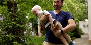 爸爸在外面和他的金发小儿子玩。抱着他，玩耍。又与他亲嘴，放在地上。绿色的草。慢动作