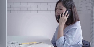 美丽的年轻微笑的亚洲女人在笔记本电脑上工作，而坐在家里的客厅。亚洲商业女性在她的家庭办公室使用电话工作。享受在家的时光。