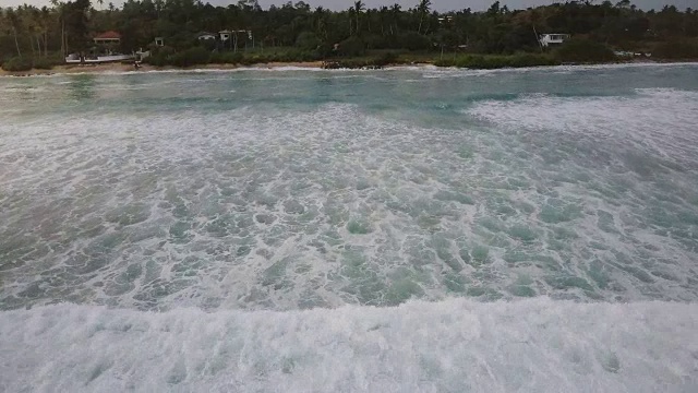 无人机摄像机跟随巨大的蓝色海洋泡沫海浪冲到美丽的异国度假胜地海岸热带树木和房屋