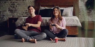 一对可爱的年轻男女正在家里卧室的地板上玩电子游戏，手握操纵杆。情感的恋人正在享受游戏。